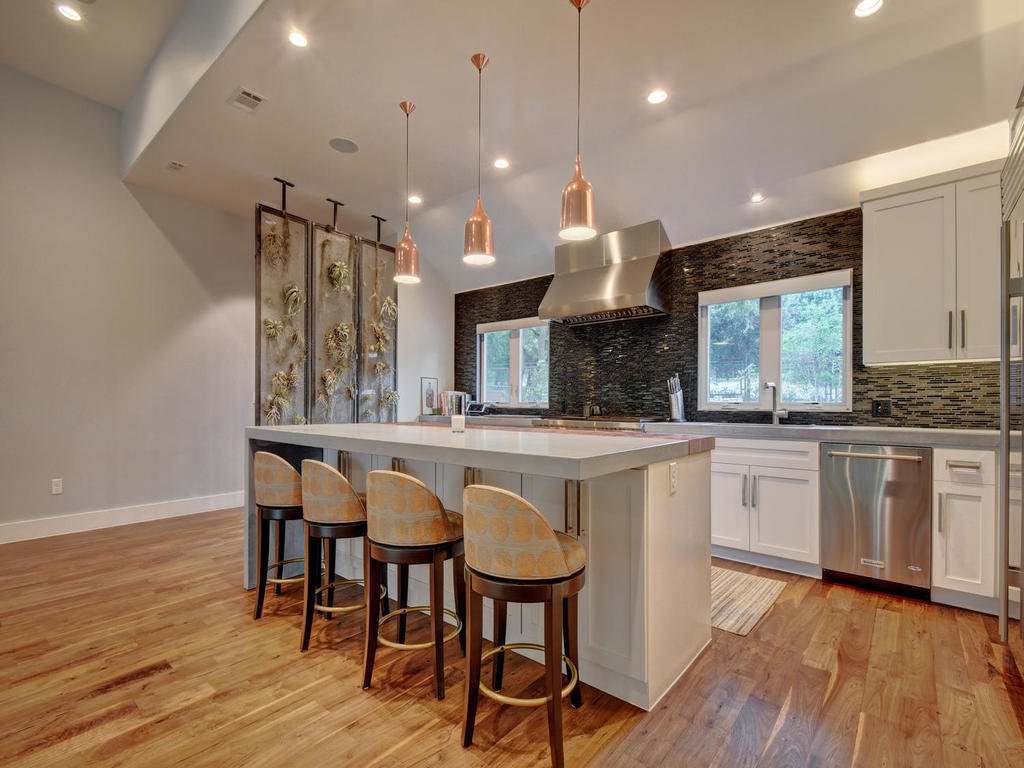 Modern kitchen with white cabinets, a large island with four bar stools, stainless steel appliances, dark backsplash, and pendant lighting.