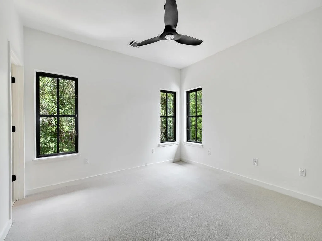 Empty white bedroom with three black-framed windows showing green trees outside, ceiling fan, white walls, and carpeted floor.