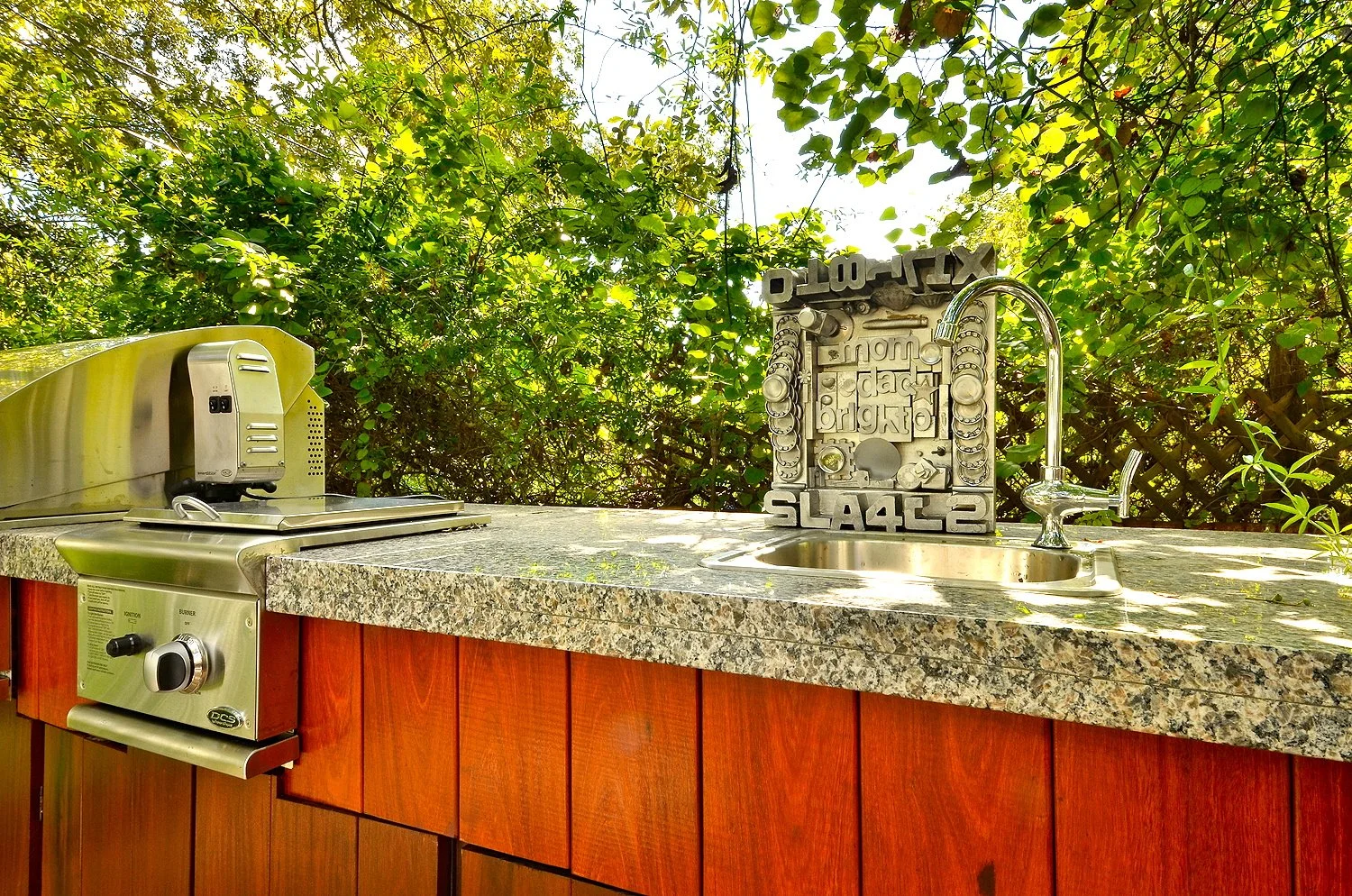 Outdoor outdoor kitchen with a stainless steel grill, a small sink with a chrome faucet, and a countertop made of speckled granite. The background is filled with lush green trees and bushes.