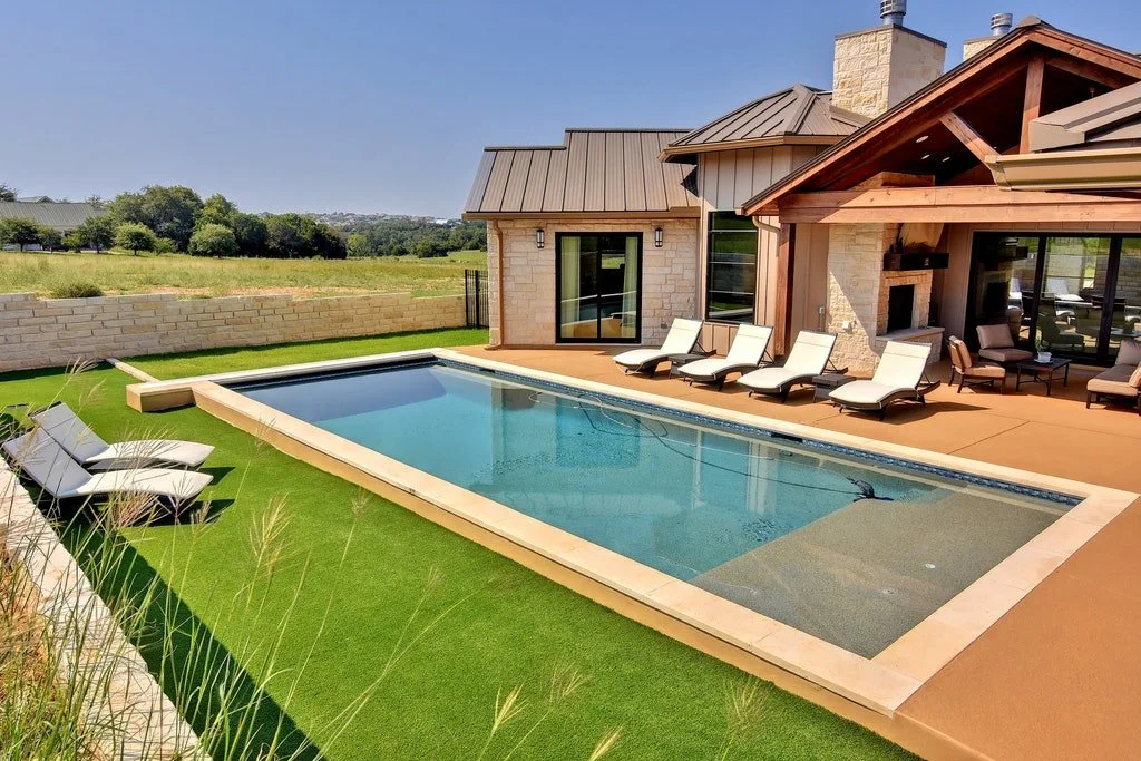 A modern backyard with a rectangular swimming pool, lounge chairs, and a patio attached to a house with large windows and a fireplace.