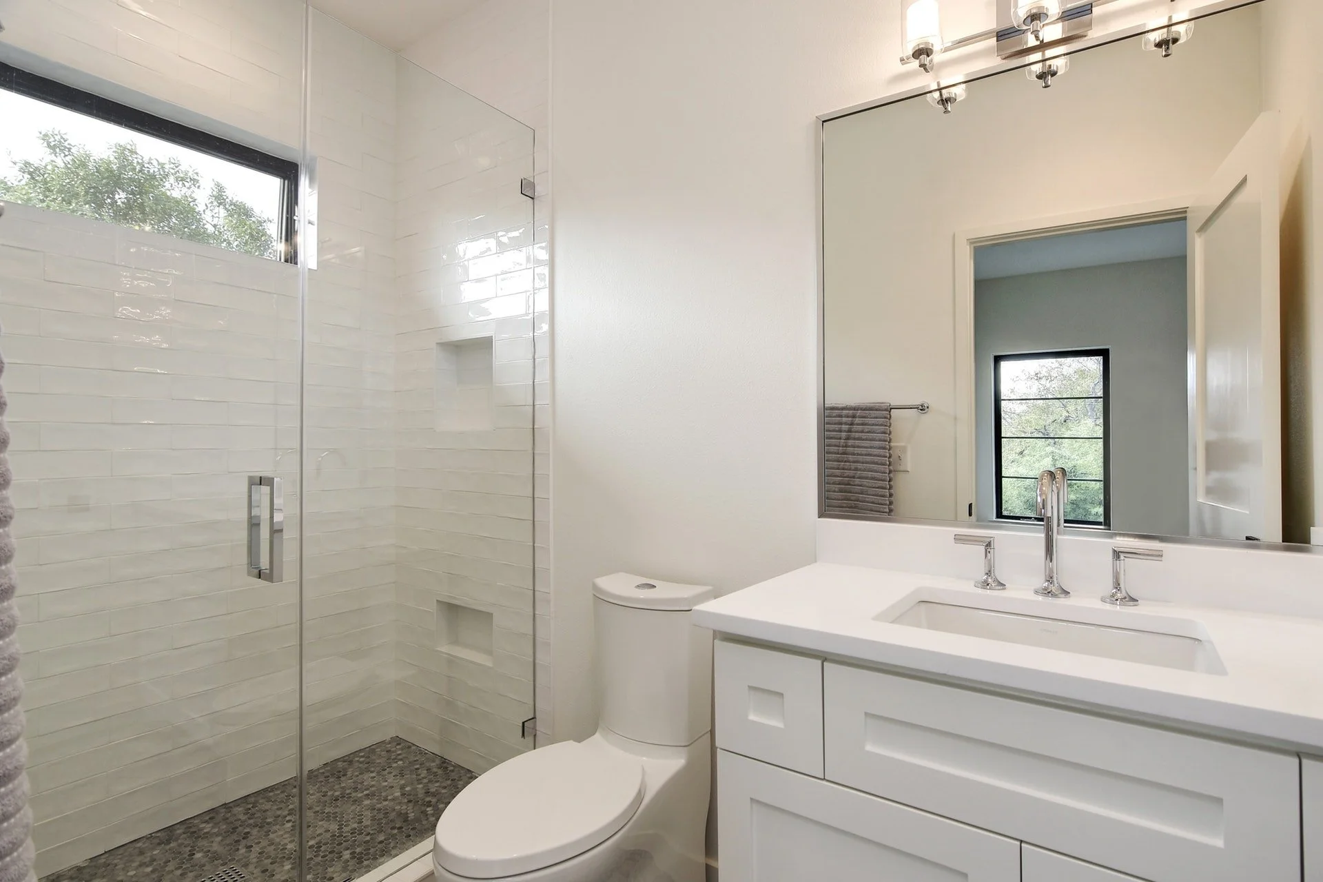 Modern bathroom with glass-enclosed shower, white walls, and a large mirror above the vanity. A window provides natural light, and there is a towel draped on a bar next to the mirror.