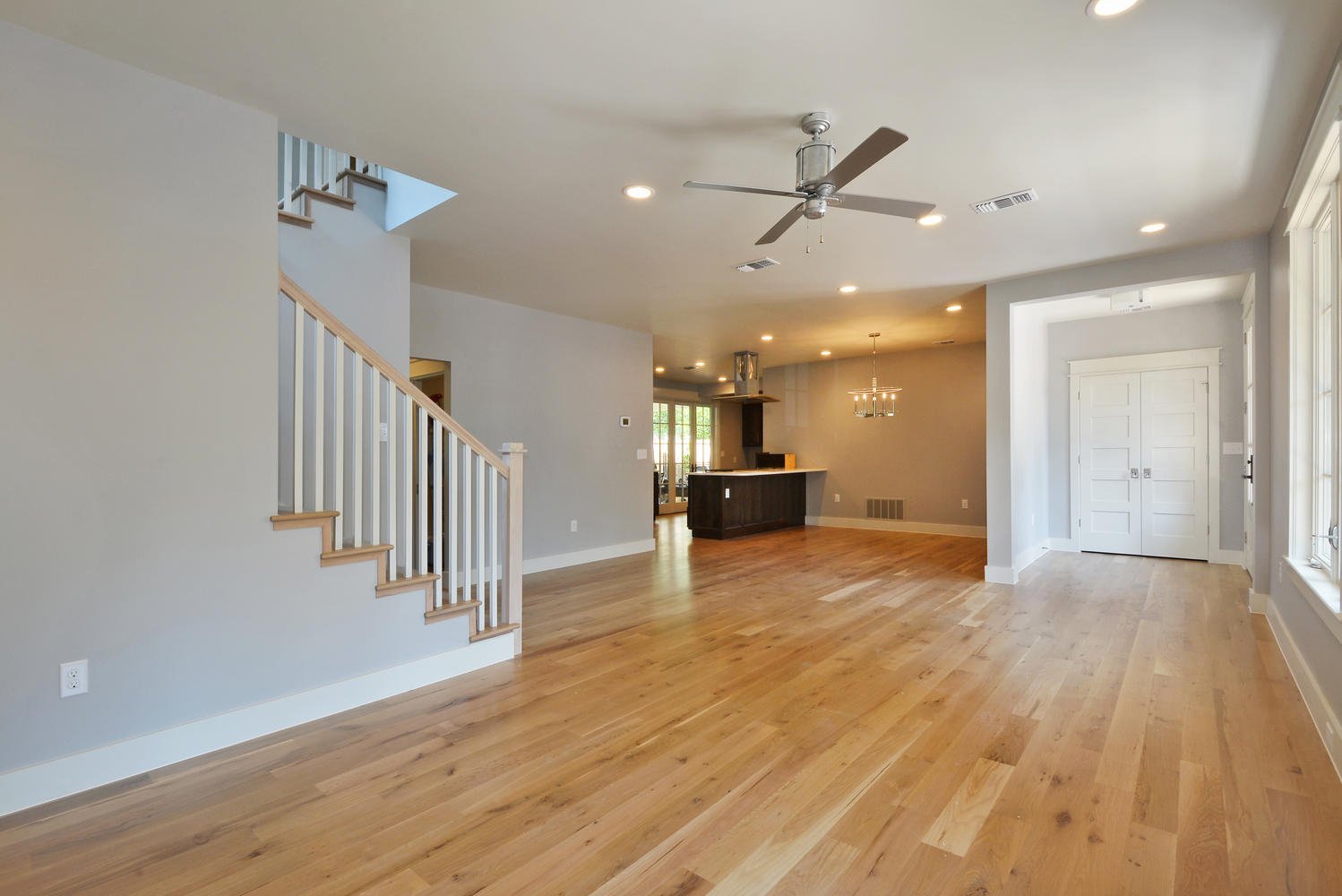 Empty living room with hardwood floors, ceiling fan, large windows, and an open kitchen area.