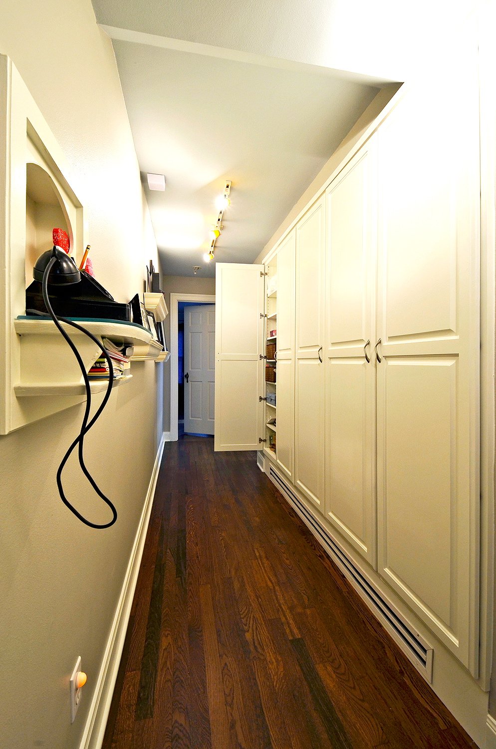 A narrow hallway with dark hardwood flooring, white walls, a long white cabinet on the right, and a small white shelf with books and electronic items on the left wall leading to a door at the end.