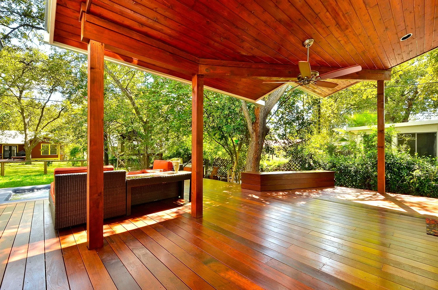 Wooden deck with outdoor furniture, ceiling fan, and surrounded by green trees and bushes.