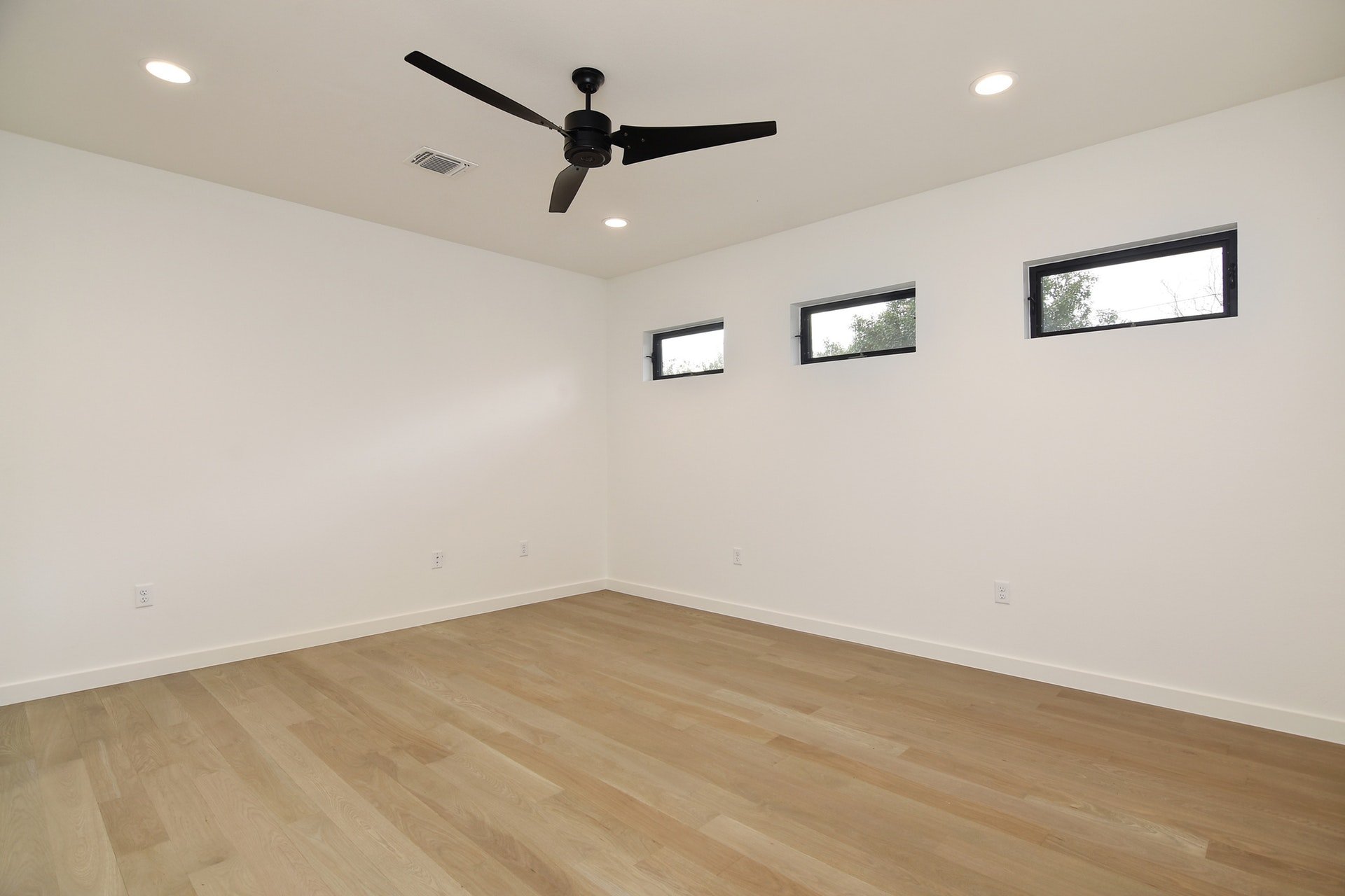 An empty room with white walls, wooden flooring, three rectangular windows near the ceiling, and a black ceiling fan.