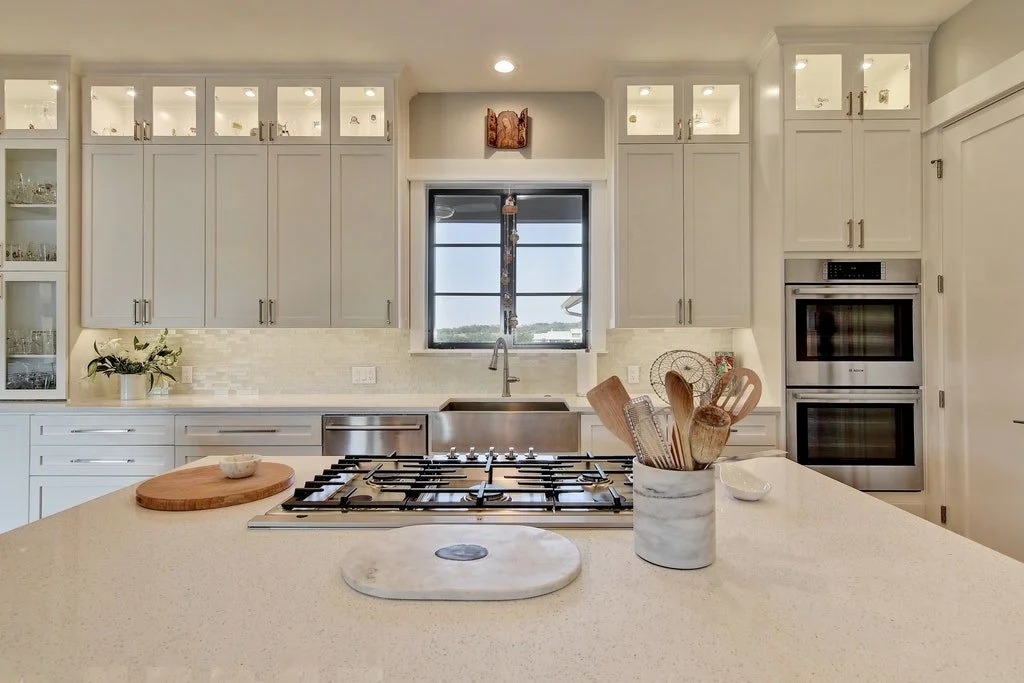 Kitchen with white cabinets, a window above the sink, a gas stove on a large island, and built-in ovens to the right. There are kitchen utensils and decorative items on the countertop.
