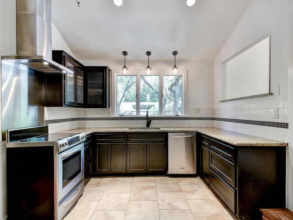 Kitchen with black cabinets, white tiled walls, granite countertops, stainless steel stove, black pendant lights, large window showing trees outside, and a white range hood.