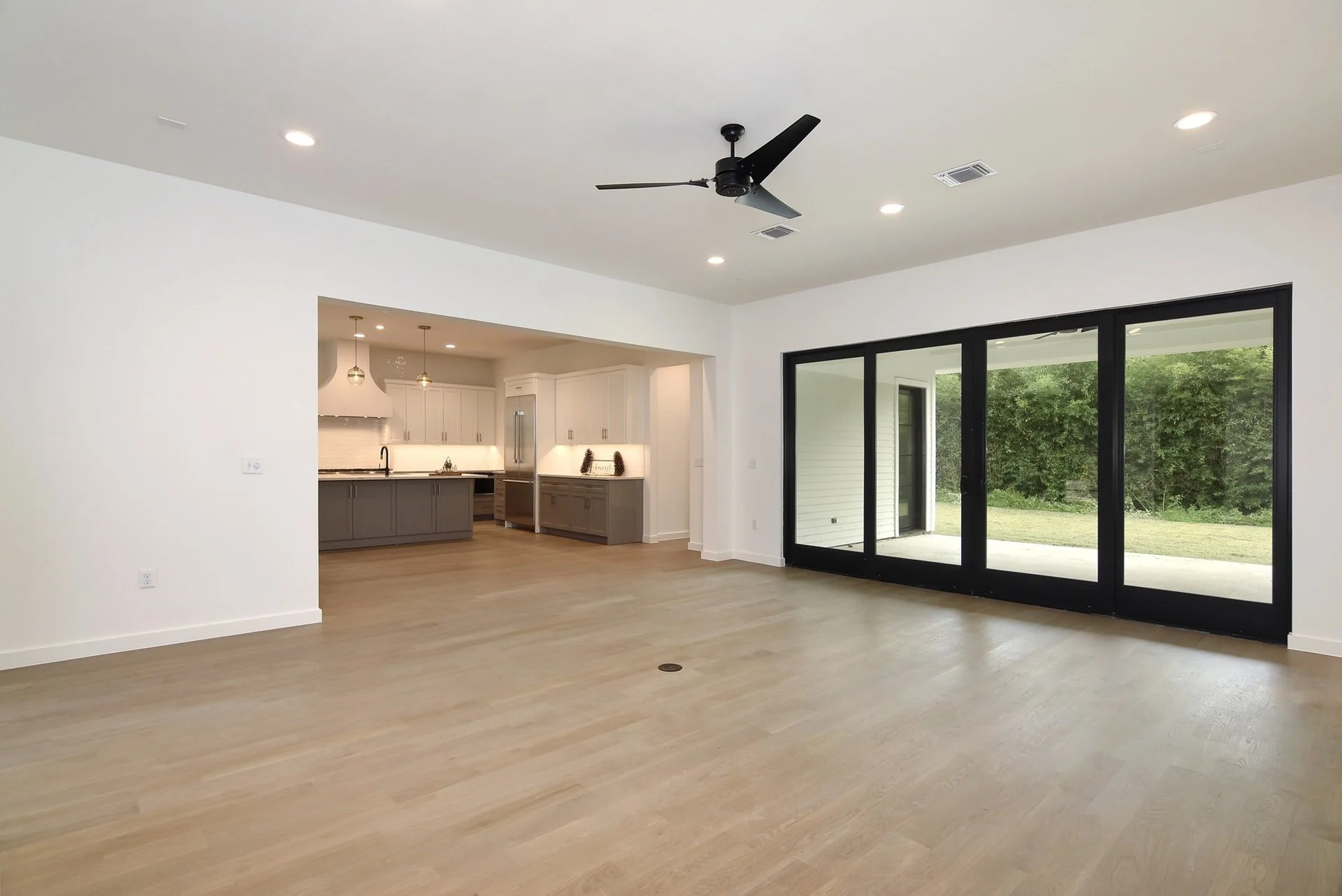 Open living room with large sliding glass doors leading outside, kitchen in background with white cabinets, gray lower cabinets, and pendant lights.