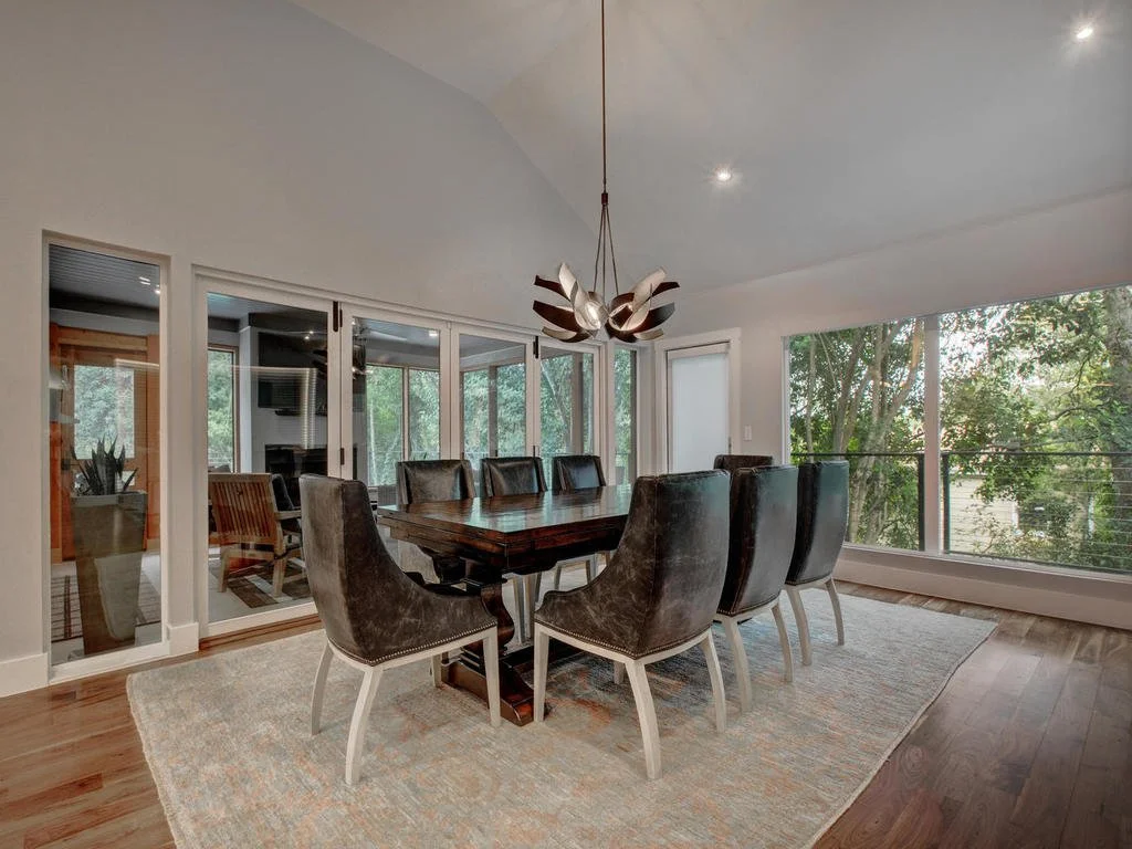A dining room with a wooden table, eight dark-colored chairs, a modern pendant light, large windows overlooking trees, and a sliding glass door leading to an outdoor deck.