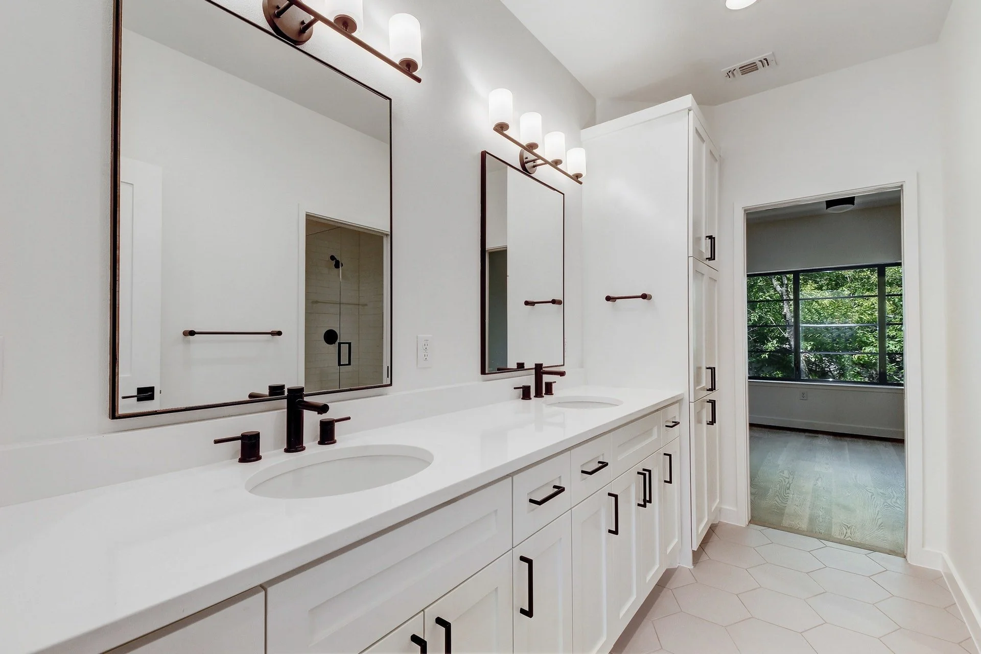 Modern bathroom with white double vanity, black fixtures, large mirrors, wall-mounted lighting, built-in cabinets, and a doorway leading to a room with a large window showing green trees.
