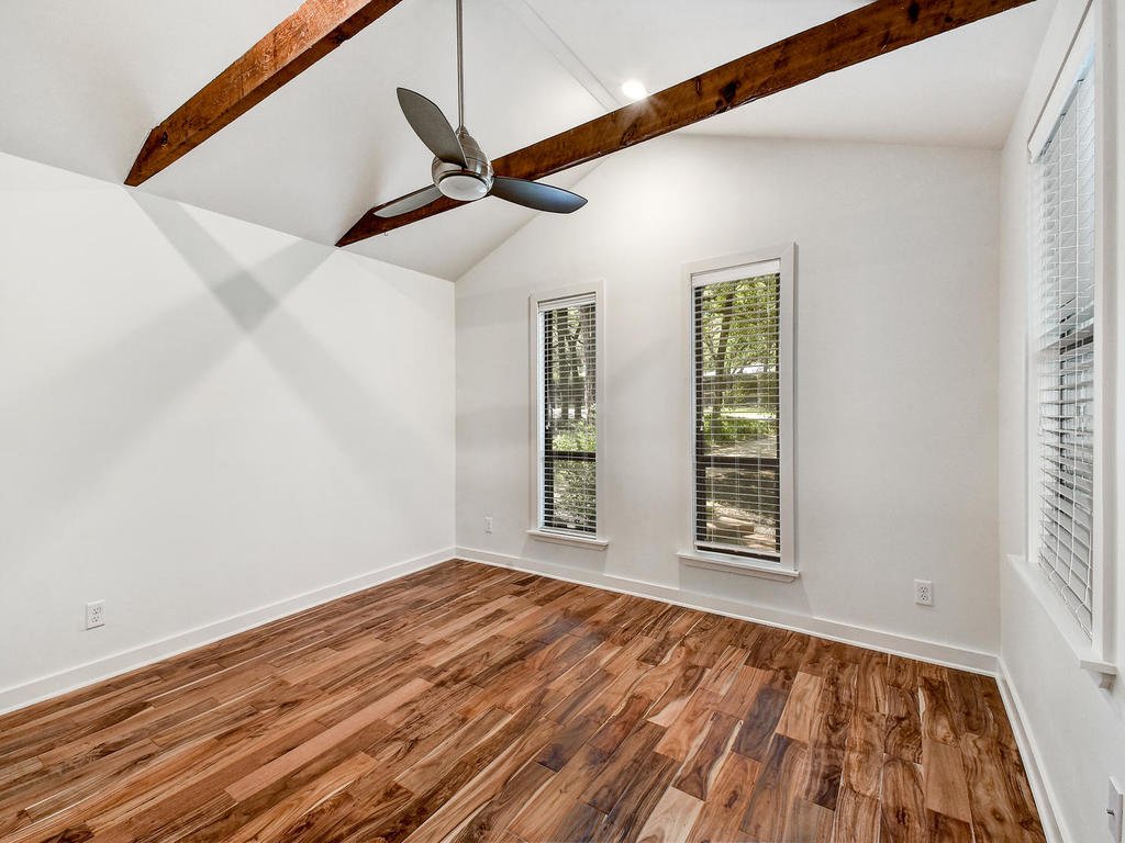 Empty room with wooden floors, white walls, three windows with blinds, a ceiling fan, and exposed wooden beams on the ceiling.