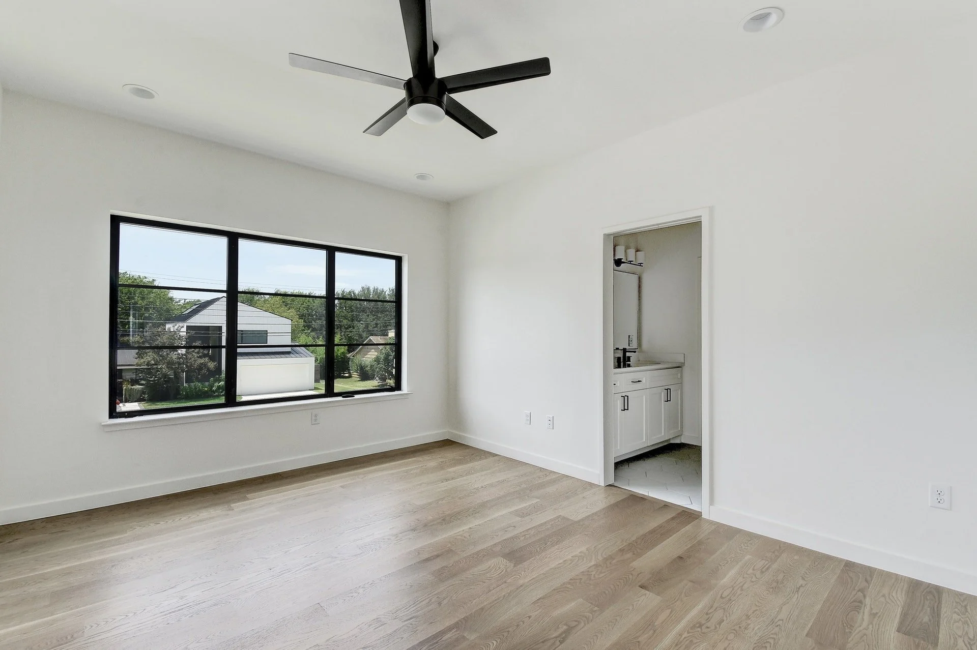 Empty living room with large window, ceiling fan, white walls, light wood flooring, and a small built-in sink area.
