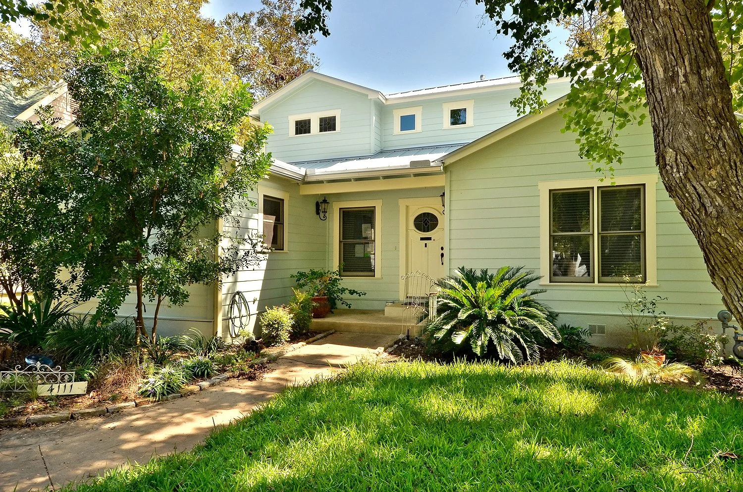 Front yard of a light green house with a small porch, surrounded by trees and bushes, with a grassy lawn and a concrete walkway leading to the door.