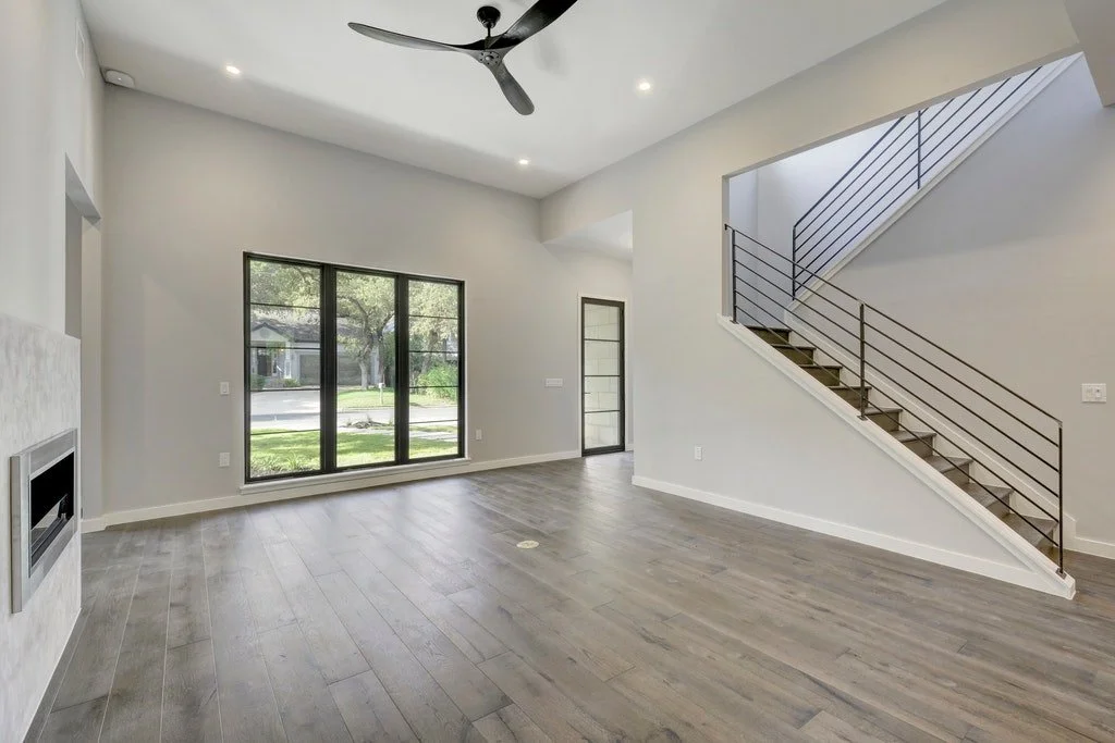 Interior view of an empty modern living room with large windows, a ceiling fan, gray flooring, a staircase with black railing, and a fireplace on the left wall.