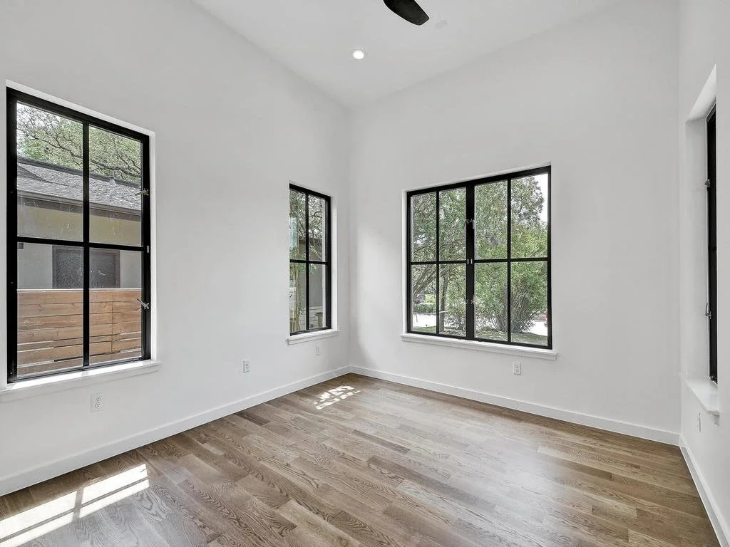 Empty room with white walls, hardwood floors, and three large black-framed windows showing outdoor trees and a neighboring house.