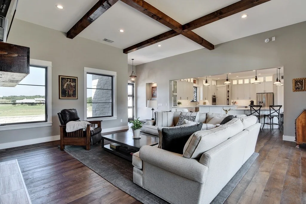 Bright living room with white walls, dark wood beams on the ceiling, large windows, a beige sectional sofa, a dark wooden coffee table, and a cozy armchair with a blanket. Opens to a modern kitchen with white cabinets and stainless steel appliances.