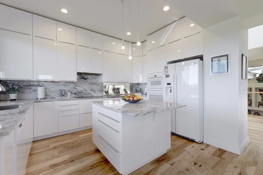 Modern white kitchen with marble countertops and island, refrigerator, microwave, and wooden flooring.