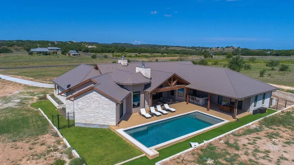 A spacious house with a swimming pool in the backyard, surrounded by a lawn and open fields, under a partly cloudy sky.