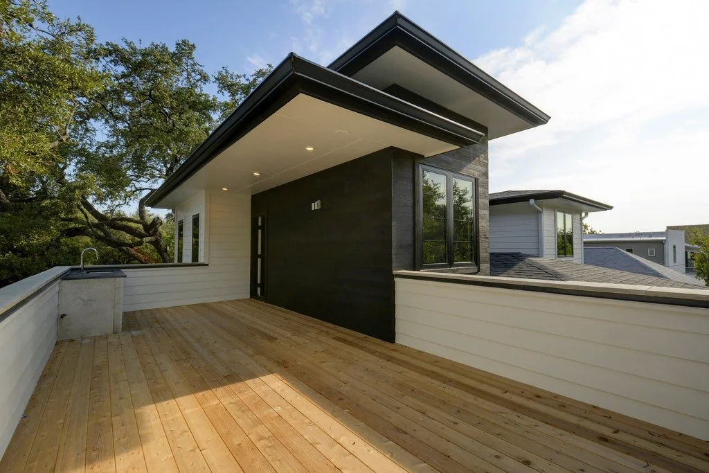 A spacious outdoor balcony with wooden flooring and white railing, attached to a modern house with black and white exterior walls, overlooking neighboring rooftops and green trees.