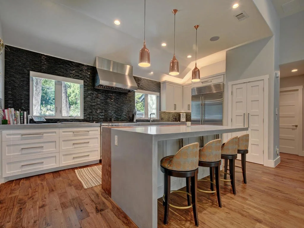 Modern kitchen with white cabinets, stainless steel appliances, black textured backsplash, and hardwood floors. Features a large island with three cushioned bar stools, pendant lights, and windows overlooking trees.