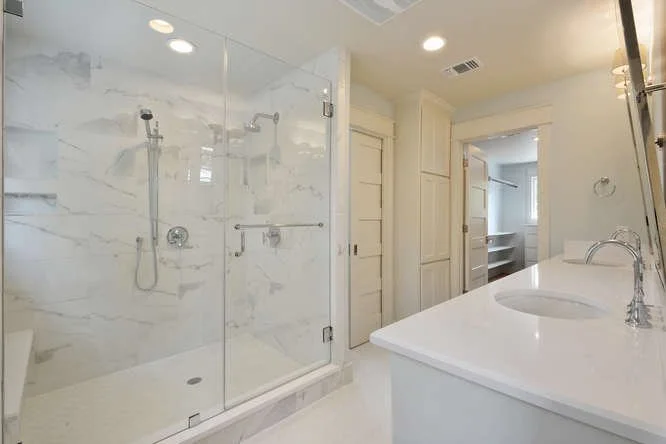 Modern bathroom with a glass-enclosed shower with marble tiles, a white vanity with a marble countertop, and a mirror. Recessed lighting and a small open shelving closet are visible.