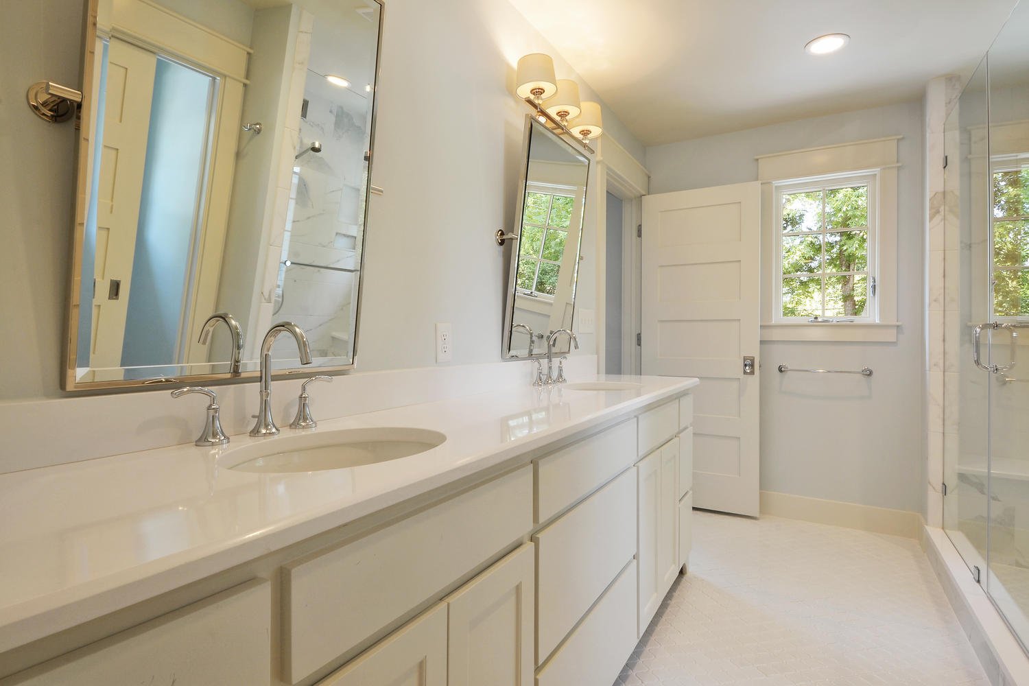 Bright bathroom with dual sinks, large mirrors, window with greenery outside, and a glass shower stall.