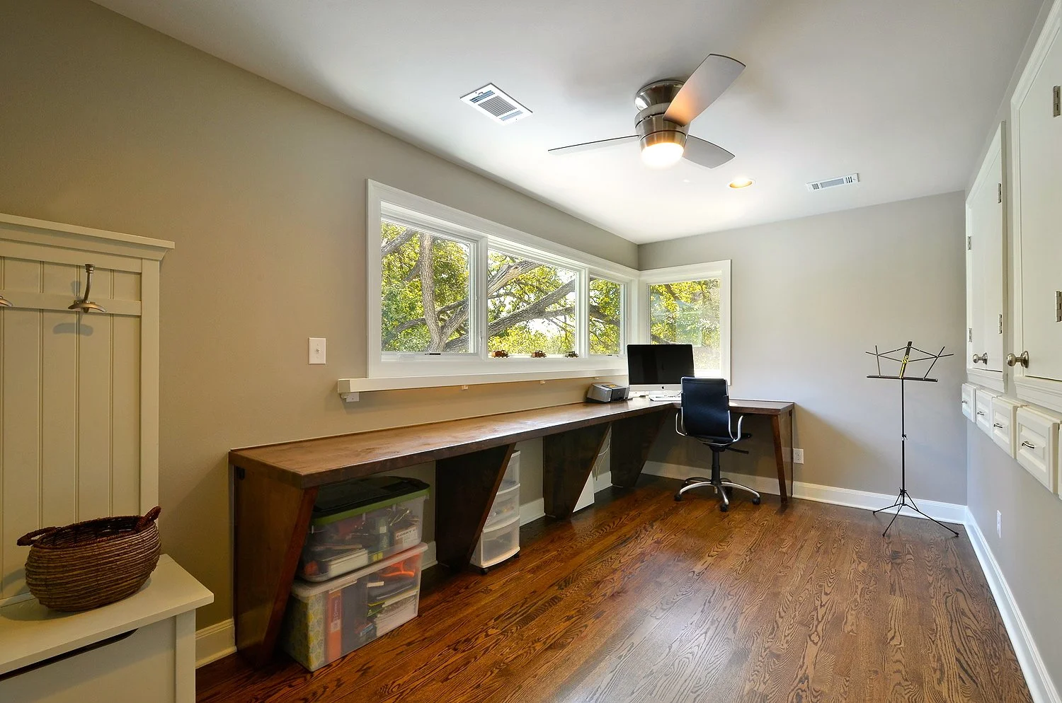 Home office with a long wooden desk near a bay window, a black office chair, a computer monitor, a music stand, and storage bins underneath the desk in a room with wooden flooring and light-colored walls.