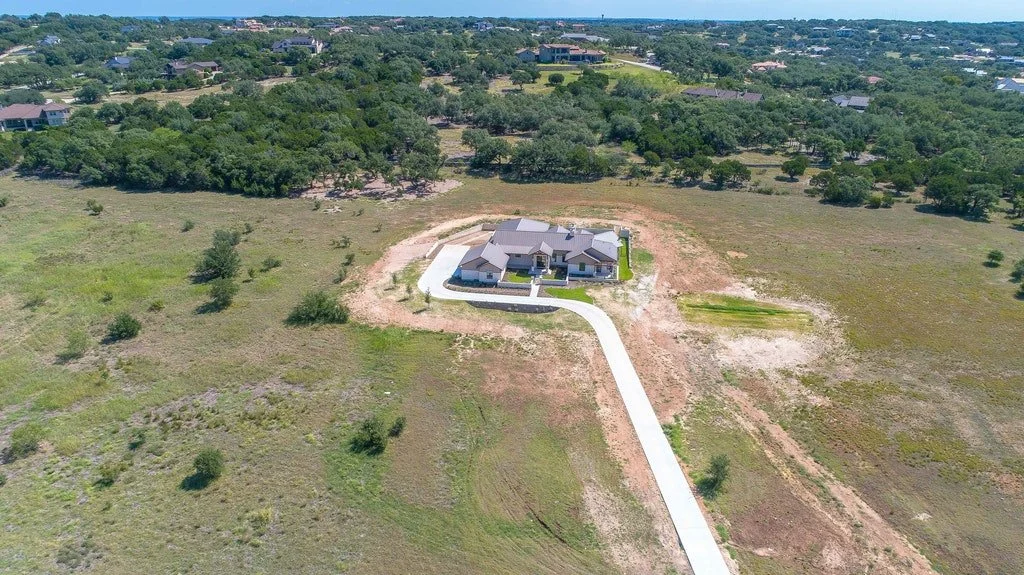 Aerial view of a newly built house with a long driveway, surrounded by open land and trees in a suburban area.