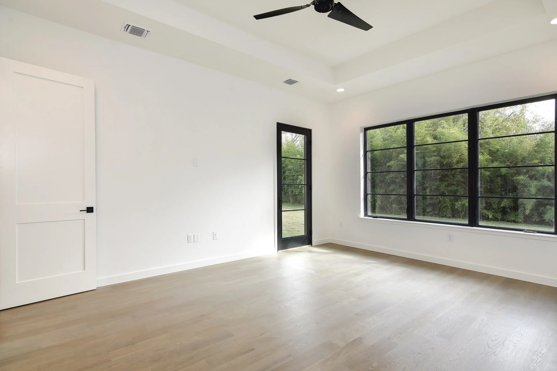 Empty room with white walls, light wood flooring, large black-framed window, black door, ceiling fan, and outside greenery visible through windows.