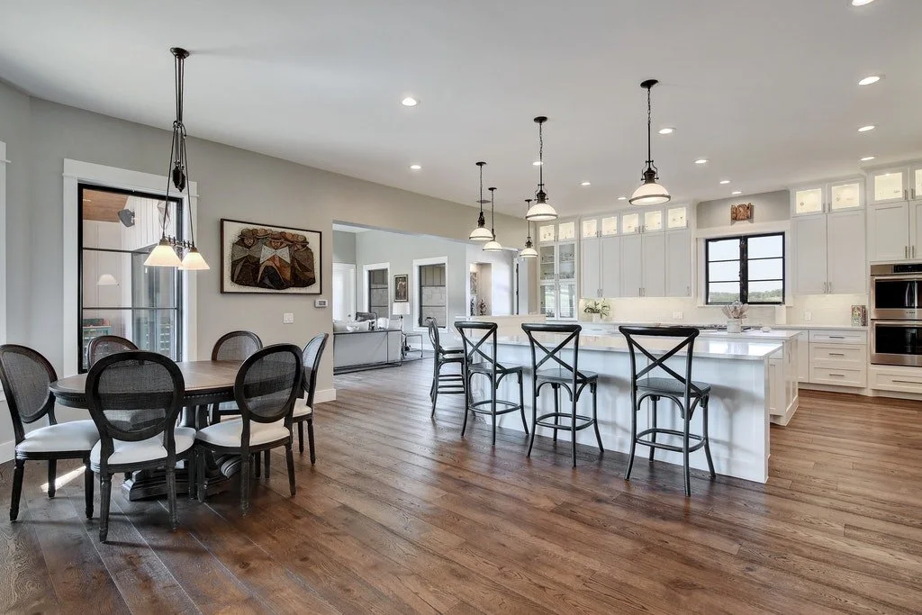 Open-concept kitchen and dining area with wooden floors, white cabinets, and a kitchen island with six black barstools. A round dining table with six chairs is on the left, and pendant lighting hangs above both areas.