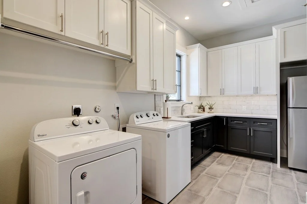 Laundry room with white and black cabinets, washer and dryer, stainless steel refrigerator, and sink under a window.
