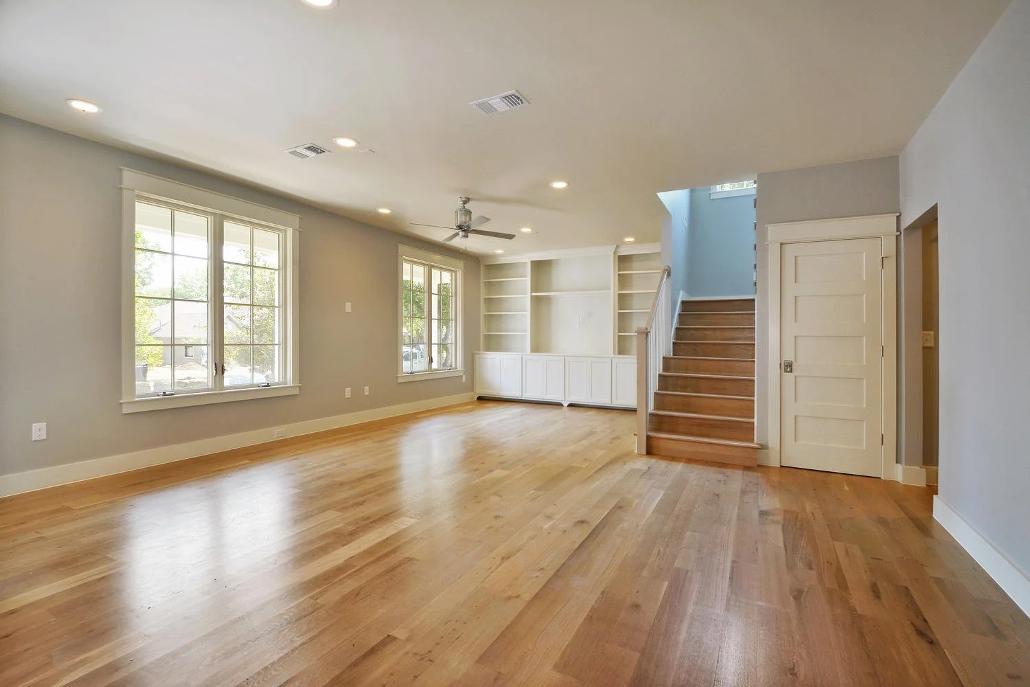 Empty living room with hardwood floors, light gray walls, large windows, built-in shelves, a ceiling fan, and a staircase.