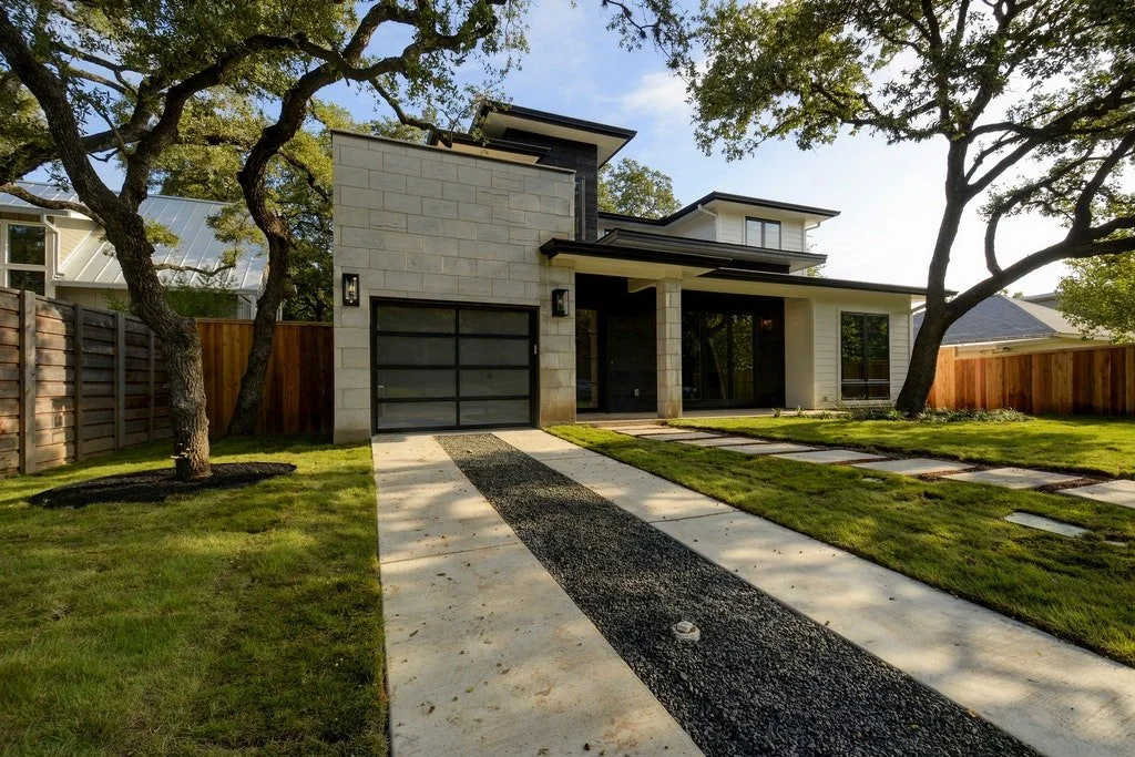 Modern house with large windows and a two-car garage, situated in a landscaped yard with trees and a stone-paved walkway.
