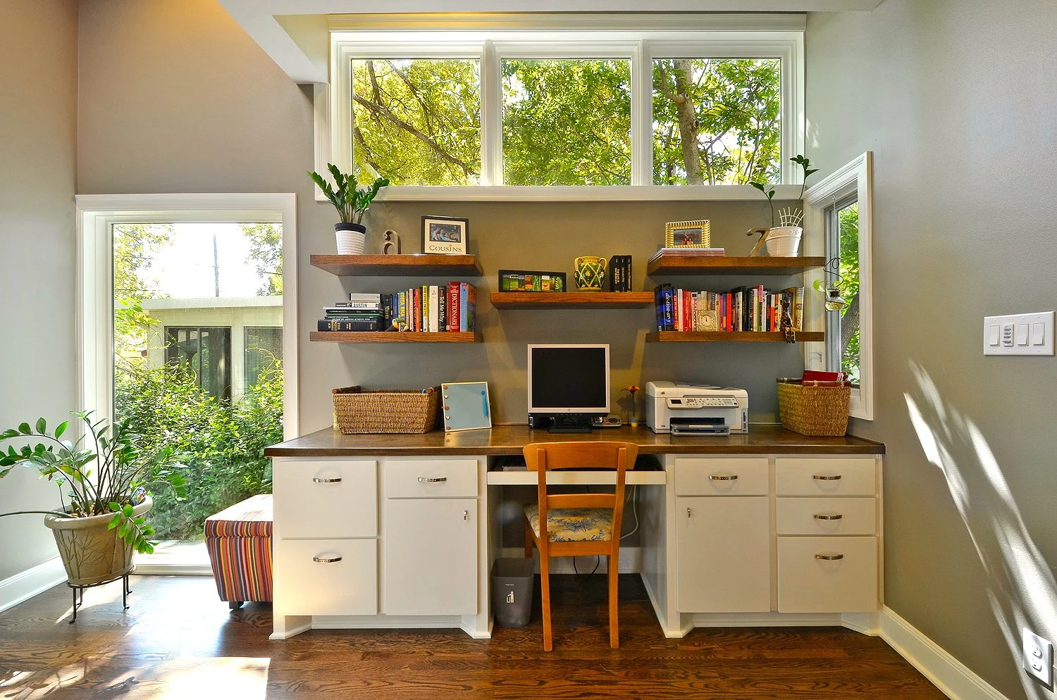 Home office with a built-in desk, white cabinets, and dark wood countertops. A computer monitor, printer, and various office supplies are on the desk. Shelves above hold books, picture frames, and decorative items. A large window above the shelves an