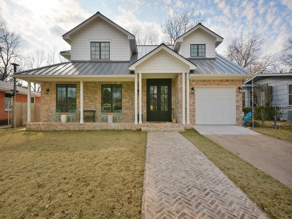 A two-story house with a brick yard and a metal roof, featuring a front porch with outdoor seating, a black front door, and an attached garage.