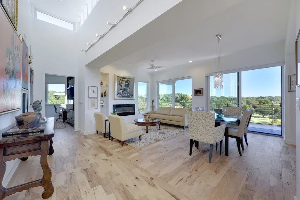 Bright living room with large windows, white sofa, armchairs, artwork, glass dining table, and balcony with outdoor view.