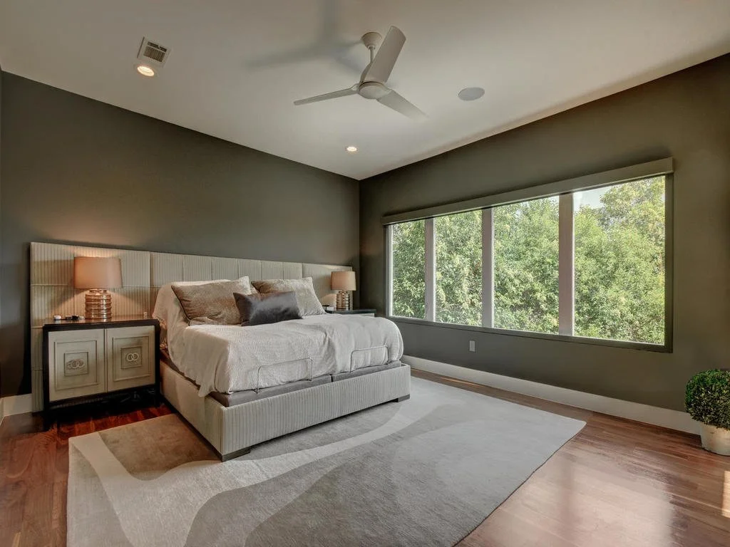 A bedroom with a large bed, nightstands with lamps, a large window showing green trees, a ceiling fan, and hardwood floors.