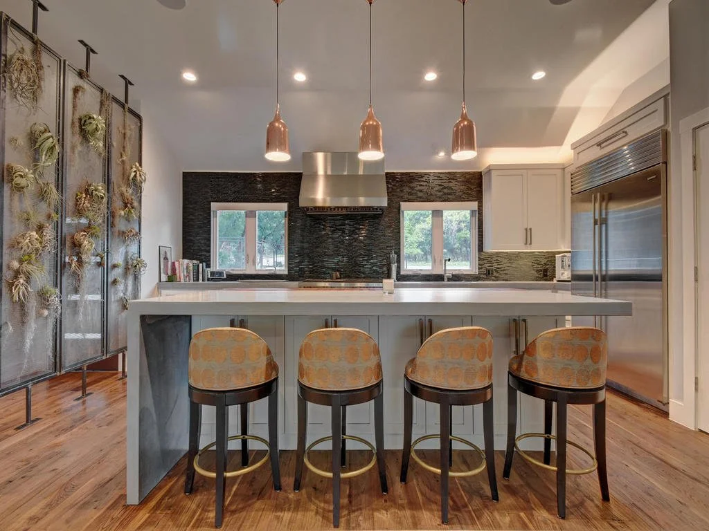 Modern kitchen with white cabinetry, dark textured backsplash, stainless steel refrigerator, and a large white island with four upholstered barstools.