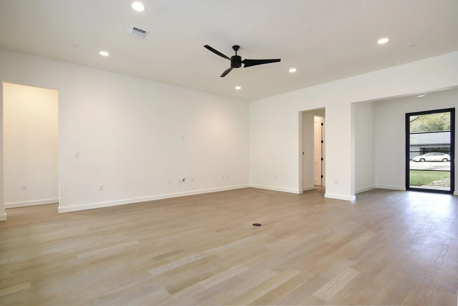 Empty living room with white walls, light wooden flooring, black ceiling fan, recessed lights, and a large glass door leading outside.