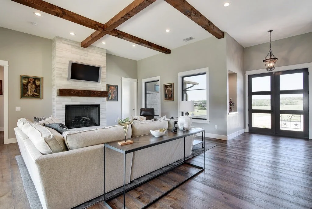 Living room with beige sofa, wooden beams on ceiling, fireplace with mounted TV, side table with lamp, hardwood floors, large window, and black front door with glass panels.