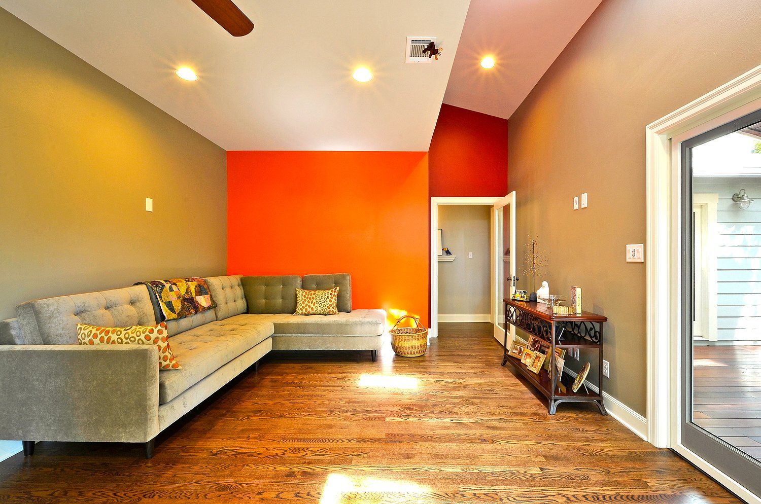 Living room with a large beige sectional sofa, a red accent wall, a wooden console table with framed photos, a sliding glass door showing an outdoor porch, and hardwood floors.