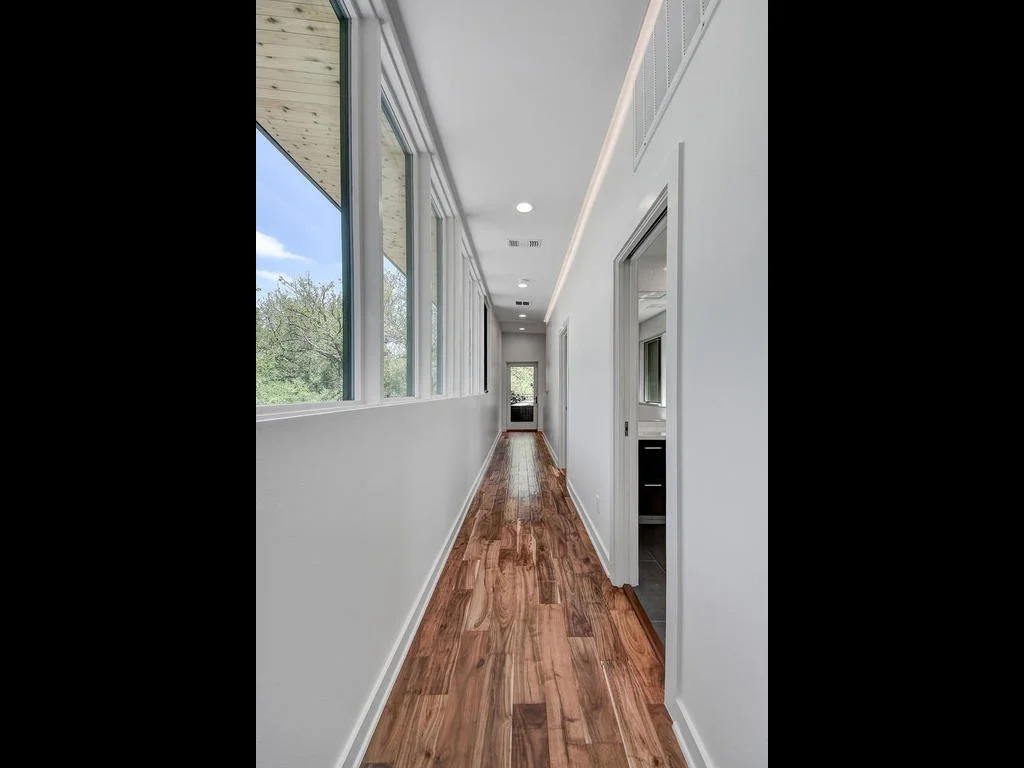 Bright hallway with large windows on the left, white walls, hardwood floor, ceiling lighting, and doors leading to other rooms.