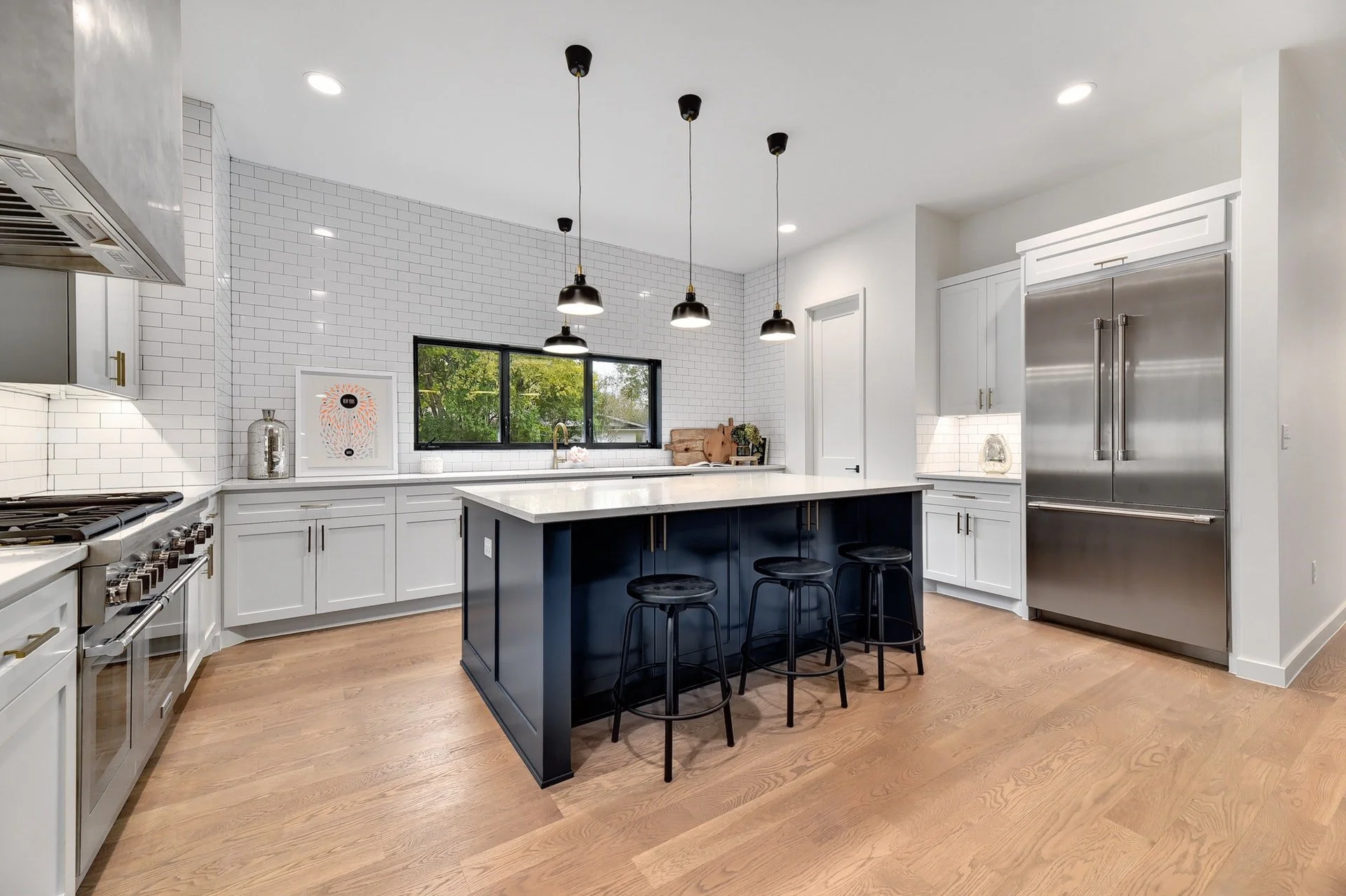 Modern kitchen with white cabinets, black island, stainless steel refrigerator, wood flooring, black pendant lights, white subway tile backsplash, and a window showing greenery outside.