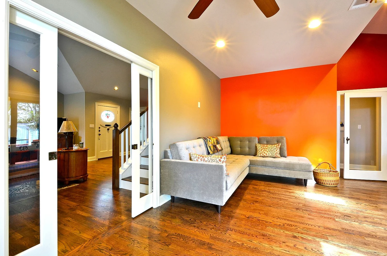 Living room with hardwood floor, orange accent wall, gray sofa with patterned pillows, basket, and ceiling fan.