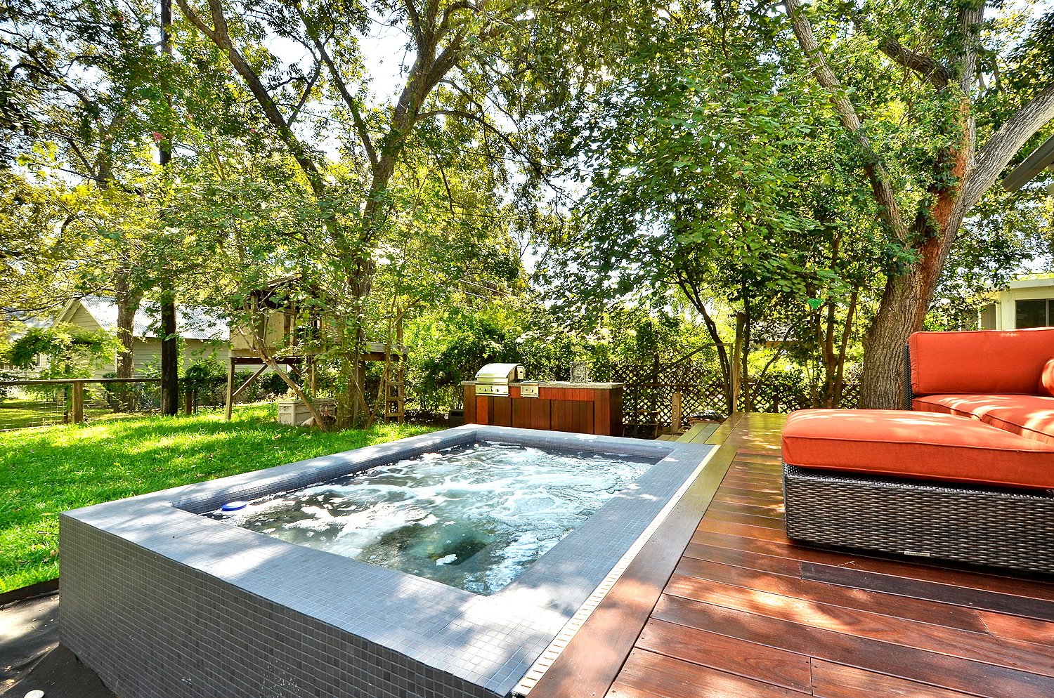 Backyard with hot tub on wooden deck, outdoor sofa, trees, and barbecue grill, sunny day