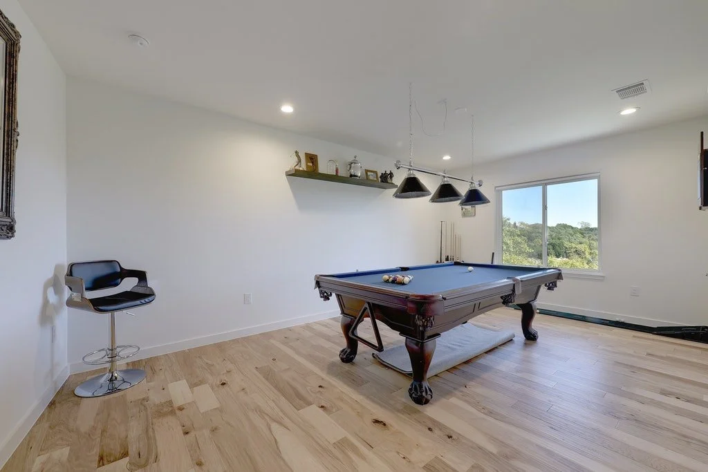 A room with a pool table, a black barstool, a wall shelf with decorative items, and a window with a scenic view of trees outside.