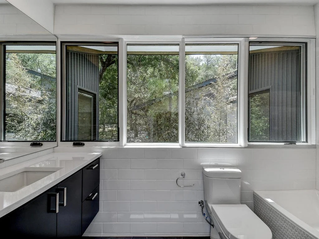 A modern bathroom with large windows overlooking trees, a white toilet, black cabinetry with a white countertop, and a bathtub.