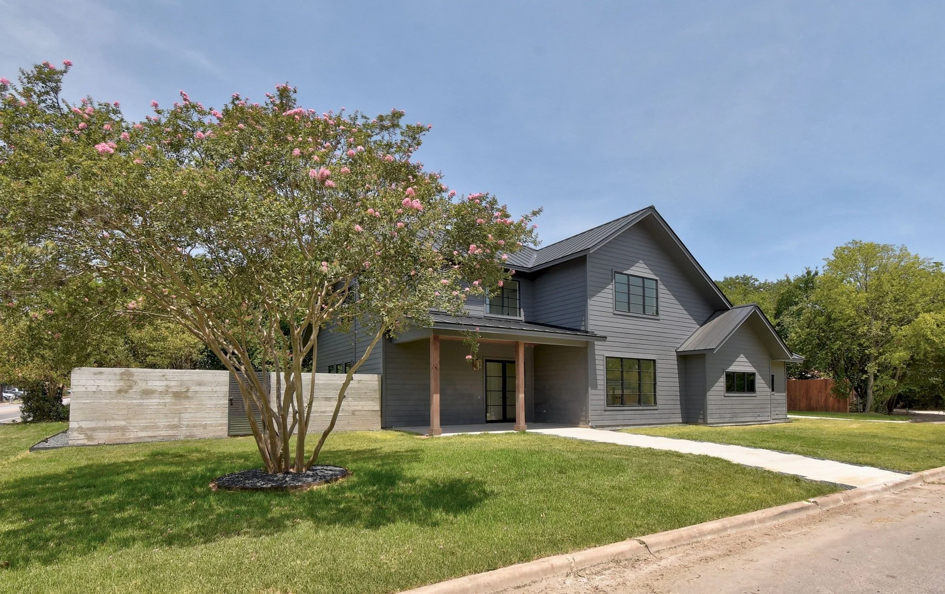Modern gray two-story house with large windows and a front porch, surrounded by a green lawn, trees, and a blue sky.
