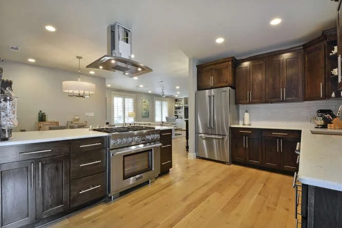Modern kitchen with dark wood cabinetry, stainless steel refrigerator, and stainless steel oven range with hood. Light hardwood floors, white walls, and recessed ceiling lights. Natural light coming through windows.