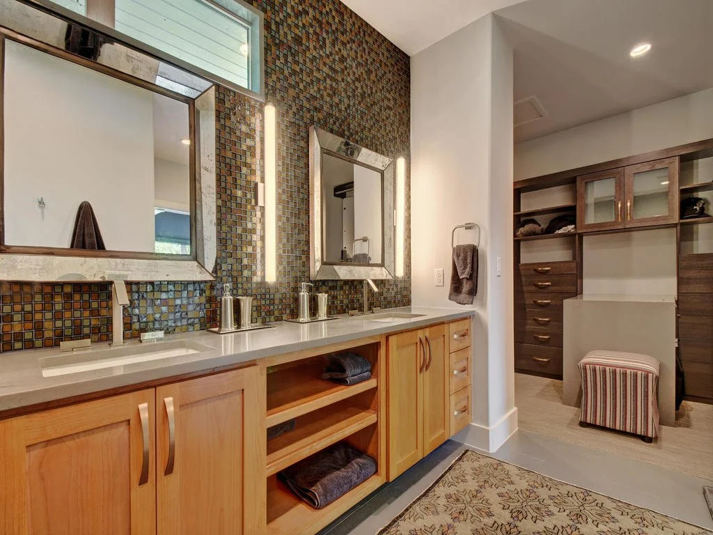 Bathroom vanity with two sinks, large mirrors, mosaic tile backsplash, light wood cabinets, and a walk-in closet with wooden shelves and a striped ottoman.